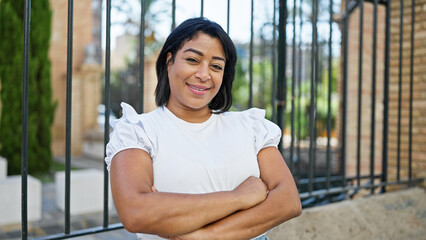 Smiling hispanic woman in white standing confidently with arms crossed in an urban park setting.
