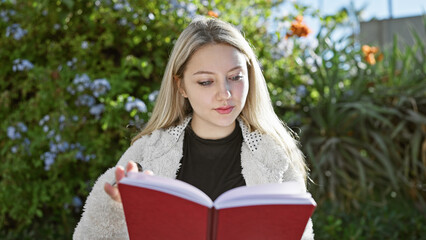 Young blonde woman reading a book in a lush garden, displaying leisure, education, and nature.
