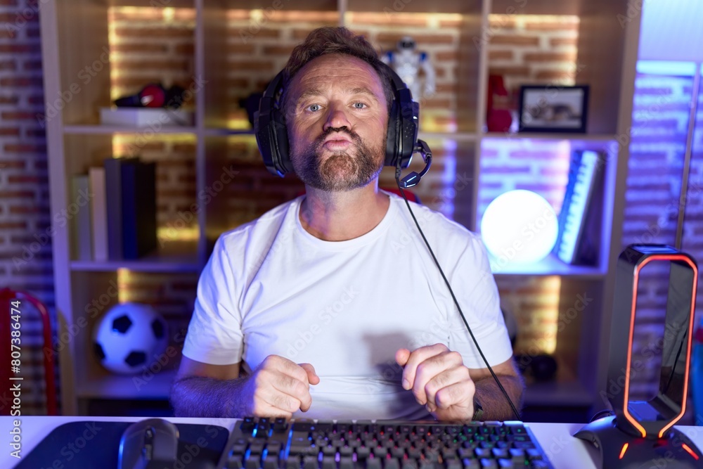 Poster Middle age man with beard playing video games wearing headphones looking at the camera blowing a kiss on air being lovely and sexy. love expression.