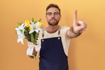 Middle age man with beard florist shop holding flowers pointing with finger up and angry...