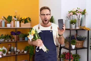 Middle age man with beard working at florist shop showing smartphone screen in shock face, looking...