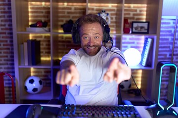 Middle age man with beard playing video games wearing headphones pointing to you and the camera with fingers, smiling positive and cheerful