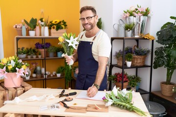 Middle age man florist holding bouquet of flowers writing on notebook at flower shop