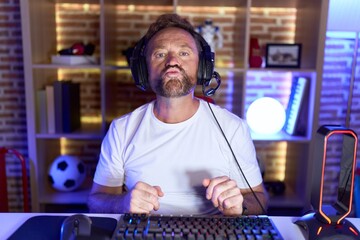 Middle age man with beard playing video games wearing headphones looking at the camera blowing a...
