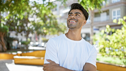 Handsome young hispanic man smiling outdoors in a city park, with trees and benches.