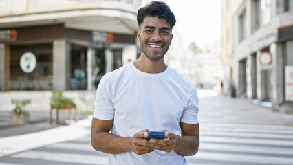 Smiling hispanic man using smartphone on sunny city street.