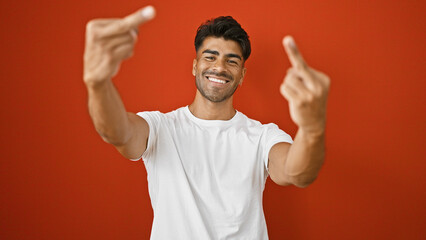 A smiling young hispanic man showing thumbs up against a plain red wall, portraying positivity.