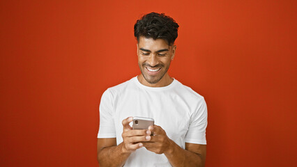 Handsome young hispanic man in white shirt smiling at phone against isolated red background