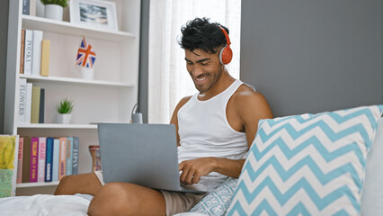 A cheerful hispanic man with beard wearing headphones enjoys using his laptop in a cozy bedroom...