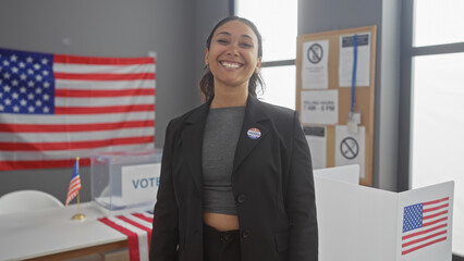 A smiling young hispanic woman with an 'i voted' sticker stands in a voting center with american...