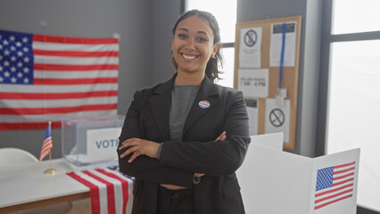 Smiling young hispanic woman with arms crossed standing in a room decorated with american flags and a voting booth indicating an election setting in the united states.
