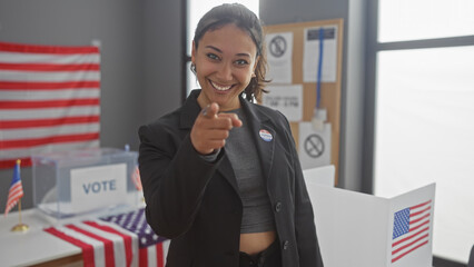 Hispanic woman pointing while smiling in an american election center with voting booths and flags.