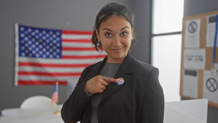 Young hispanic woman pointing to 'i voted' sticker indoors with american flag backdrop, evoking civic duty and pride.