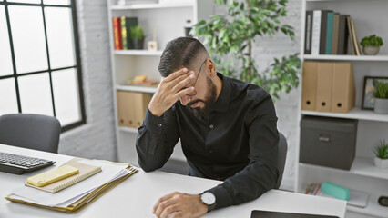 A stressed man in an office setting, with hand on head, feeling overwhelmed by work.