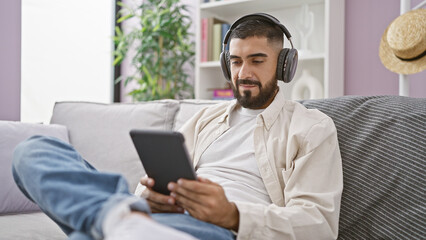 Handsome bearded man in casual wear using tablet and headphones while lounging on a sofa indoors.