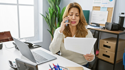 Concerned hispanic woman multitasking in an office using phone and reading document.