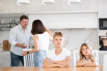 Brother and sister sitting at table in kitchen during conflict between their parents