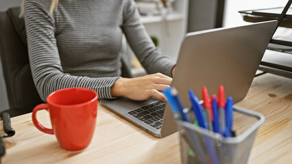 Caucasian woman typing on a laptop in a modern office, highlighted by a red mug and stationery on...