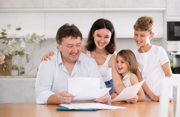 Fototapeta premium Joyful adult man examining documents while sitting at table in kitchen with his family