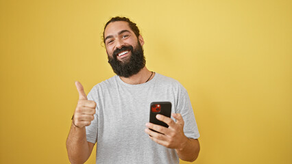 A cheerful bearded man gives a thumbs-up while holding a smartphone against a yellow background.