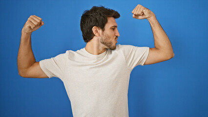 A young hispanic man flexes muscles confidently against an isolated blue background, embodying strength and vitality.