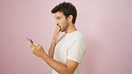 A surprised hispanic man with a beard looks at his phone against a pink background.