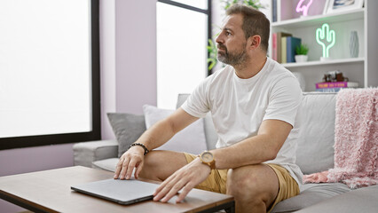 Mature handsome hispanic man sitting thoughtfully in a modern living room with a laptop.