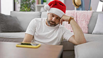 Handsome middle-aged hispanic man in santa hat showing fatigue while sitting on a living room sofa.