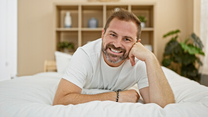 Smiling middle-aged man with grey hair lying in a minimalist bedroom at home, portraying a relaxed and comfortable atmosphere.