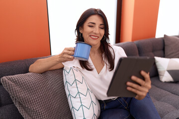 Middle age hispanic woman using touchpad drinking coffee sitting on sofa at home