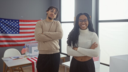 A man and a woman with arms crossed stand smiling in an indoor american electoral setting.