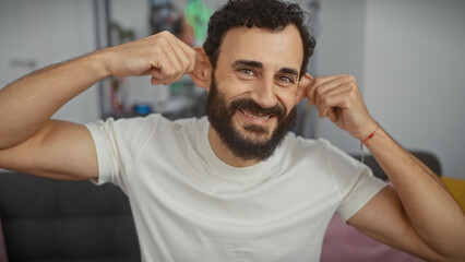Smiling bearded man playfully stretches his ears indoors at home, showcasing a casual and joyful...