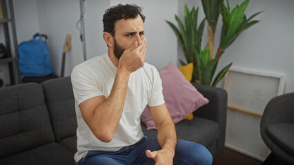 Man holding nose due to bad smell in modern living room, showing discomfort while seated on gray sofa. - obrazy, fototapety, plakaty