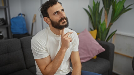 Thoughtful bearded man in casual attire sits in a modern living room, pondering with a hand on chin.