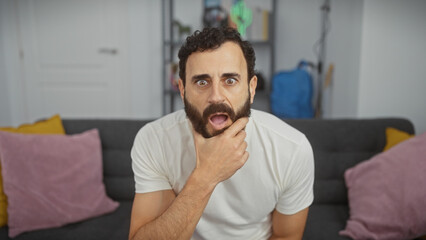 A surprised middle-aged man with a beard sitting in a modern living room, showing an expressive reaction.