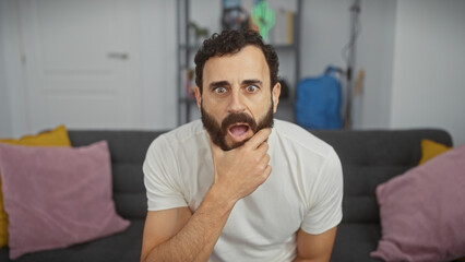 Fototapeta premium A surprised middle-aged man with a beard sitting in a modern living room, showing an expressive reaction.
