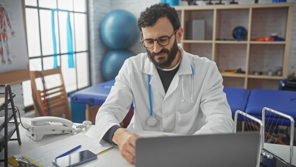A middle-aged bearded man in a white lab coat working at a laptop in a medical office setting...