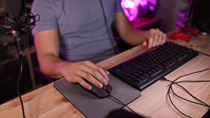 A young hispanic man uses a computer mouse and keyboard in a dark gaming room with purple led lights