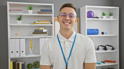 Handsome hispanic man smiling confidently at a clinic with lanyard, glasses and a bookshelf background.