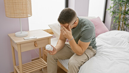 A distressed hispanic man sits on a bed holding a medicine bottle in a modern bedroom.