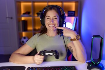 Beautiful brunette woman playing video games wearing headphones smiling cheerful showing and pointing with fingers teeth and mouth. dental health concept.