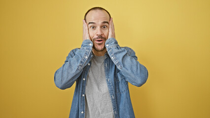 Surprised bald hispanic man with beard wearing denim expressing shock against a yellow isolated...