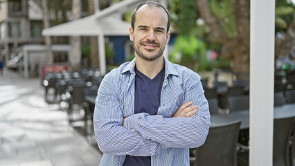 A bearded, bald hispanic man crosses his arms while standing on a city street with an outdoor cafe in the background