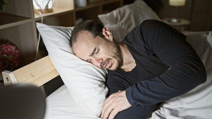 A bald hispanic man with a beard appears distressed, lying in a bedroom at night.