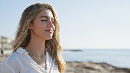 Young, beautiful blonde lady taking a calming breath with closed eyes, standing at seaside,...