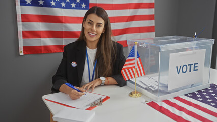 A smiling young hispanic woman sits indoors with an american flag, taking notes at an electoral...