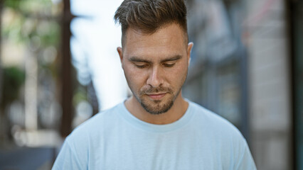 Close-up of a pensive young hispanic man with a beard, standing on a city street with a blurred...