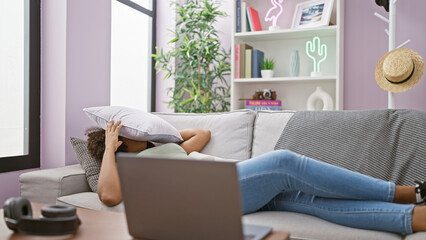 Stressed black woman with braids lying on sofa covering face with cushion indoors