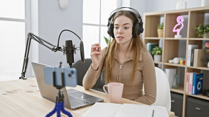 A young blonde woman records a podcast in a modern radio studio, wearing headphones and using a...