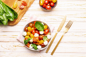 Fresh juicy homemade caprese salad with baby mozzarella cheese, cherry tomatoes, basil leaves and olive oil on white wooden table with colden cutlery set and napkin. Traditional Italian cuisine.